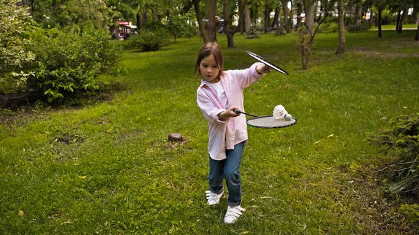 funny kid in casual clothing playing badminton on green grass in park - Foto, afbeelding
