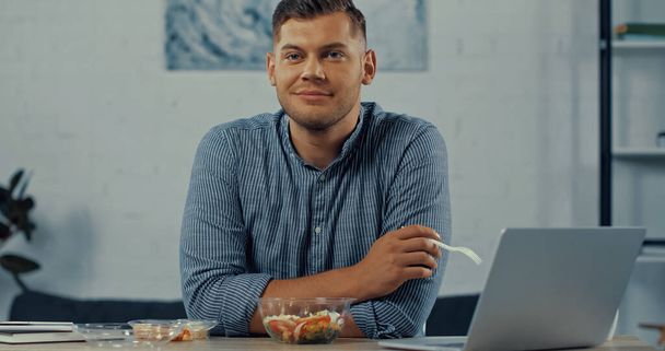 happy man holding plastic fork near salad in container and laptop on desk  - Fotó, kép