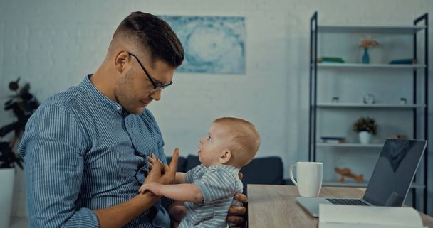 side view of father in glasses looking at infant boy while working from home  - Photo, Image