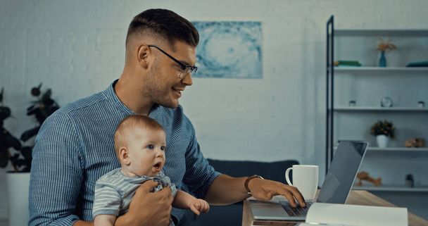 cheerful father in glasses using laptop while infant boy and working from home  - Valokuva, kuva