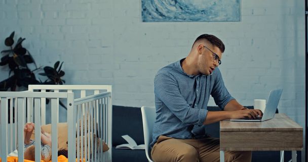freelancer in glasses talking on smartphone and using laptop while sitting near baby crib with infant son  - Fotoğraf, Görsel