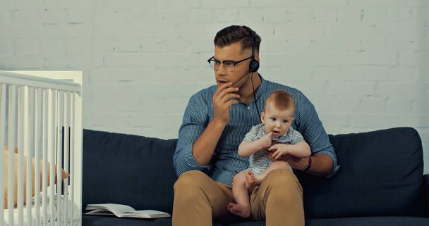 man in headset with microphone holding infant son while working from home  - Foto, Imagen