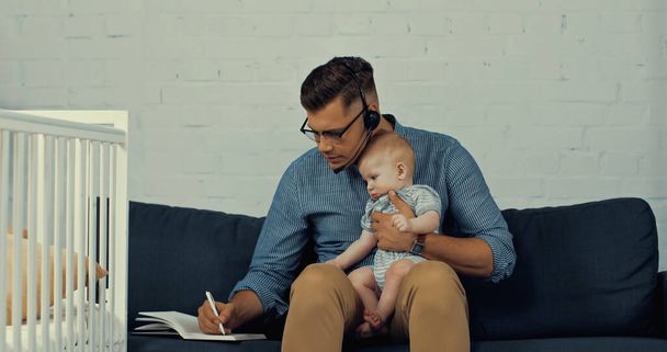 мужчина в гарнитуре с микрофоном держит в руках маленького сына, пишет в тетрадке и работает из дома  - Фото, изображение
