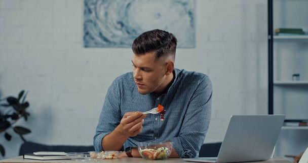 freelancer holding plastic fork and eating salad near laptop on desk - Fotó, kép