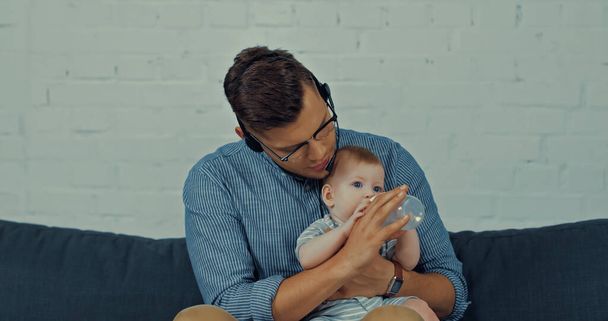 работник в наушниках с микрофоном кормящий младенца с молоком в детской бутылочке  - Фото, изображение