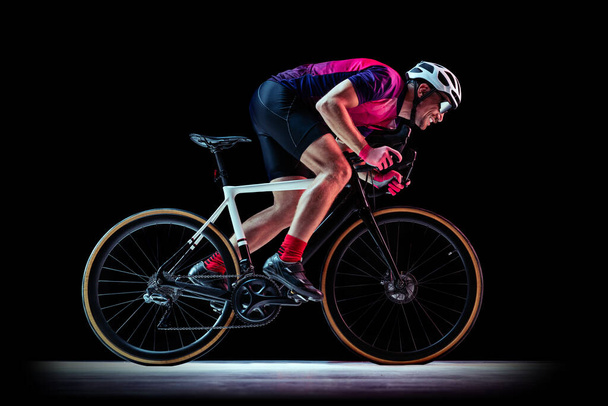 Portrait of man, professional cyclist training, riding isolated over black studio background in neon light. Concept of sport, action, motion, speed, hobby, lifestyle. Copy space for ad - 写真・画像