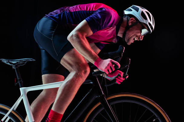 Retrato de homem, treinamento de ciclista profissional, montando isolado sobre fundo estúdio preto em luz de néon. Velocidade. Conceito de esporte, ação, movimento, velocidade, hobby, estilo de vida. Espaço de cópia para anúncio - Foto, Imagem