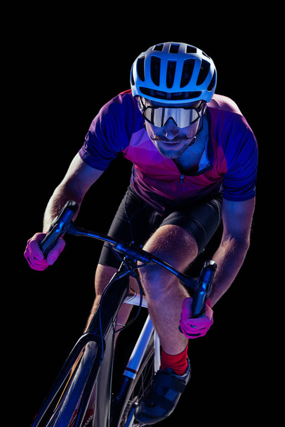 Portrait of man, professional cyclist training, riding isolated over black studio background in neon light. High speed. Concept of sport, action, motion, speed, hobby, lifestyle. Copy space for ad - Foto, Imagem