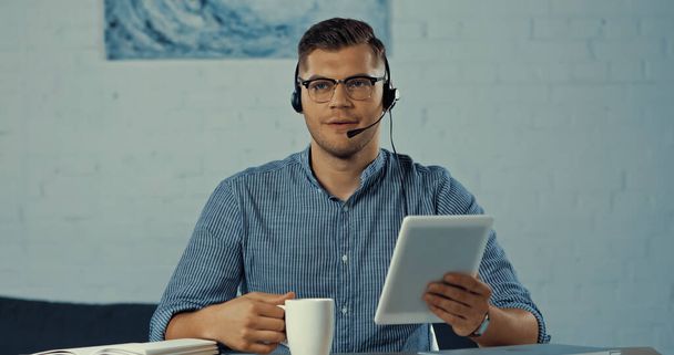 operator in headset and glasses holding cup of coffee and using digital tablet  - Foto, afbeelding