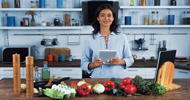 вегетаріанка з цифровим планшетом посміхається на камеру біля сирих інгредієнтів на кухні
 - Фото, зображення