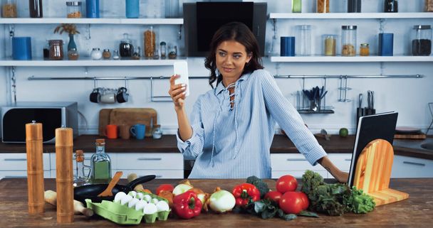 Glückliche Frau macht Selfie mit Smartphone in der Nähe roher Zutaten auf Küchentisch - Foto, Bild