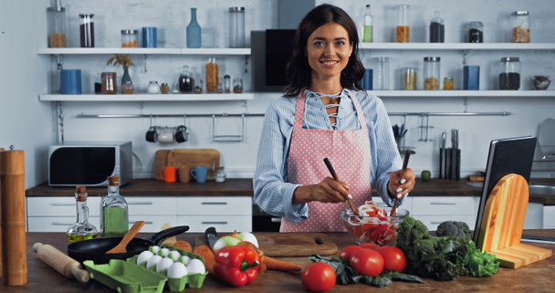 cheerful woman mixing fresh vegetable salad and smiling at camera in kitchen - Photo, image