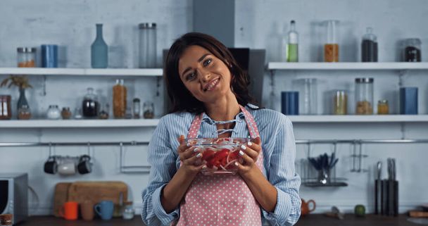 vegetarian woman in apron holding bowl of vegetable salad and smiling at camera - Fotó, kép