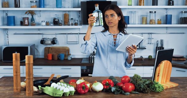 housewife with digital tablet looking at bottle with oil near raw ingredients in kitchen - Φωτογραφία, εικόνα