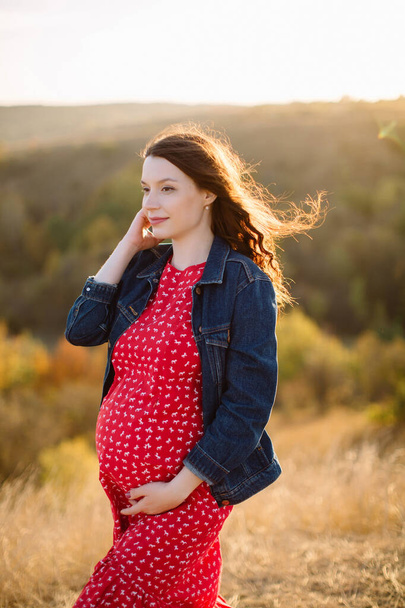 Nuori raskaana oleva äiti punaisessa mekossa pitää vatsaansa ja seisoo keskellä syksyn maisemaa kaupungin ulkopuolella. Onnellinen valmistautuminen synnytykseen, tuleva äitiys. Laadukas laukaus. - Valokuva, kuva