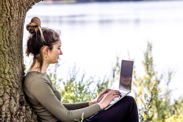 Work from anywhere. Remote freelancer work in nature using renewable energy via a foldable solar panel. Young woman, female freelancer working with laptop with Beautiful view of forest and lake - Photo, image
