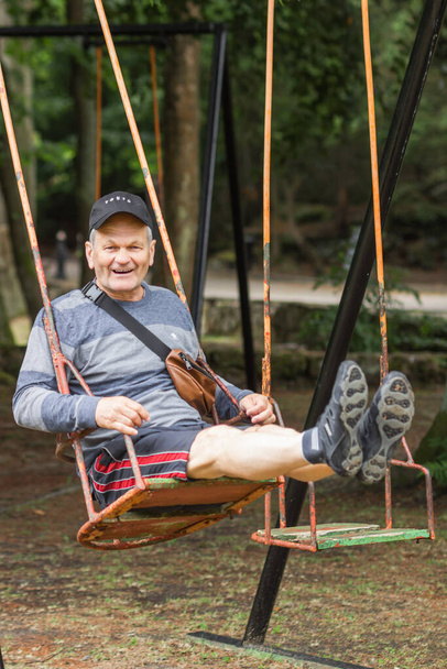 Carefree senior man in casual clothes and black cap having fun and swinging on swing in amusement park. Old man is young at heart. Active seniors and anti age concept.  - Zdjęcie, obraz