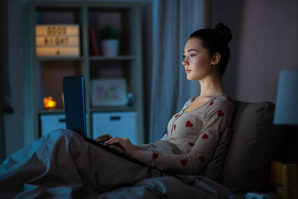 technology, bedtime and people concept - teenage girl with laptop computer sitting in bed at home at night - Fotografie, Obrázek