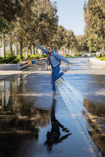 Une femme en tenue de denim danse debout sur un orteil sur l'asphalte humide d'une allée du parc. Reflet miroir dans une flaque d'eau. La joie printanière après la pluie - Photo, image