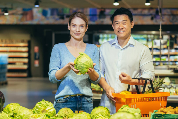 Young married couple, Asian man and blonde woman in supermarket, shoppers choose vegetables in vegetable section, portrait of happy shoppers, smiling looking at camera - Zdjęcie, obraz