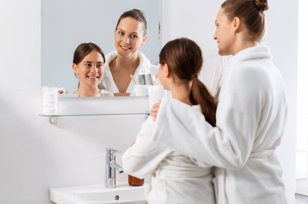 beauty, hygiene, morning and people concept - happy smiling mother and daughter looking to mirror at bathroom - Фото, изображение