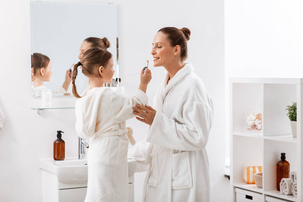 beauty, family and people concept - happy smiling mother and little daughter with lipstick applying make up in bathroom - Foto, imagen
