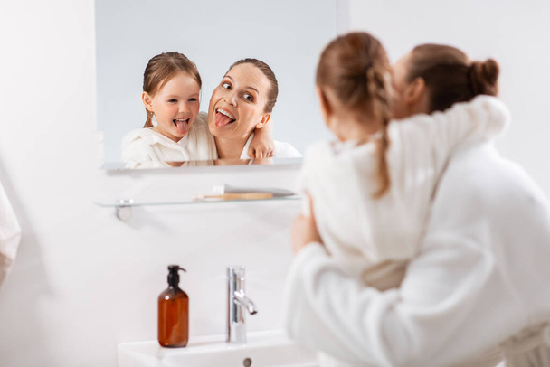 beauty, family and people concept - happy smiling mother and little daughter looking to mirror and hugging in bathroom - Photo, image