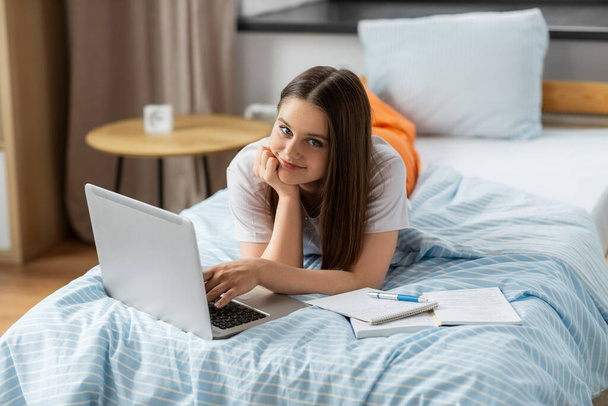 school, online education and e-learning concept - happy smiling teenage student girl with laptop computer writing to notebook lying on bed at home - Φωτογραφία, εικόνα