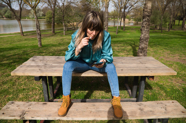 Una giovane, bella, bionda donna è seduta a un tavolo di legno nel parco, a guardare il suo cellulare e fumare una sigaretta. Sullo sfondo si può vedere il fiume in una giornata di sole. Smetti di fumare - Foto, immagini