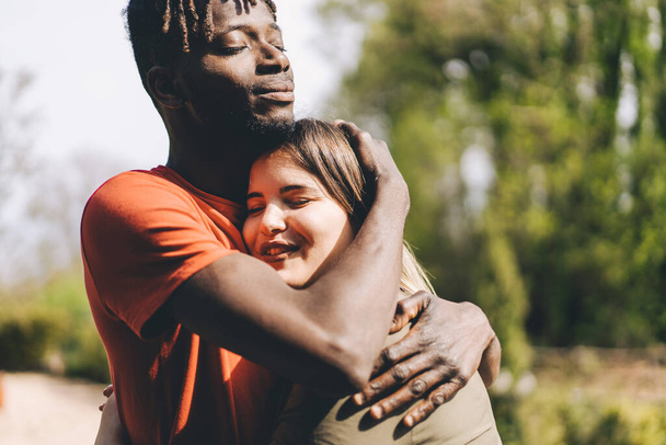 Мультирасовая пара с молодым африканцем и белой девушкой, обнимающими друг друга на открытом воздухе в парке. Концепция нежной романтики и межрасовых отношений. - Фото, изображение