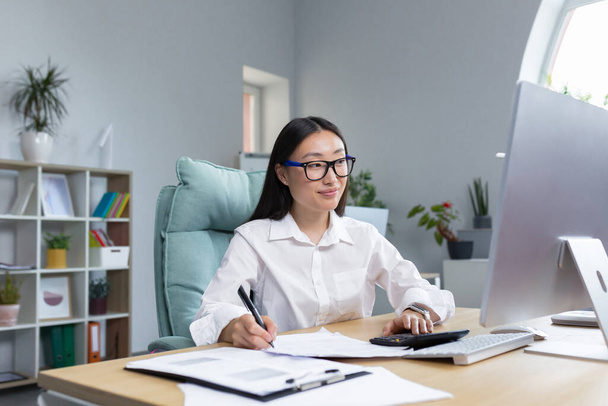 Work with documents. Portrait of a young beautiful business woman Asian accountant works with documents and reports. Sitting at the desk in the office, writing. - 写真・画像