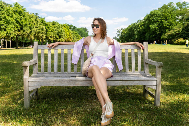 Portrait young adult attractive woman enjoy sitting on bench and relaxing calm carefree rest in city park against green grass and trees on sunny day. Single female person relaxing chilling outdoors. - Foto, afbeelding
