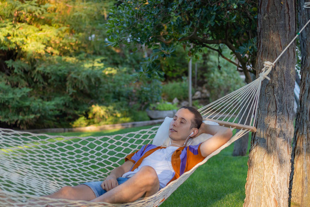 Молодий чоловік у відпустці лежить в гамаку, слухаючи музику спокійно і розслаблено з природою на задньому плані влітку
 - Фото, зображення
