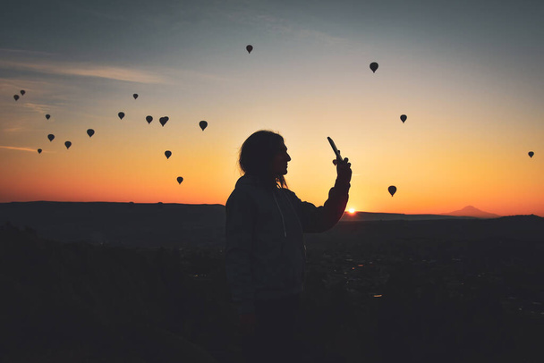 Silhouette of Smart phone in hands of woman taking pictures of a beautiful landscape and balloons in Cappadocia. Sunrise time, dreamy travel concept - Photo, image
