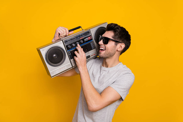 Photo of funky brunet millennial guy hold look boombox wear t-shirt glasses isolated on yellow background. - Foto, imagen