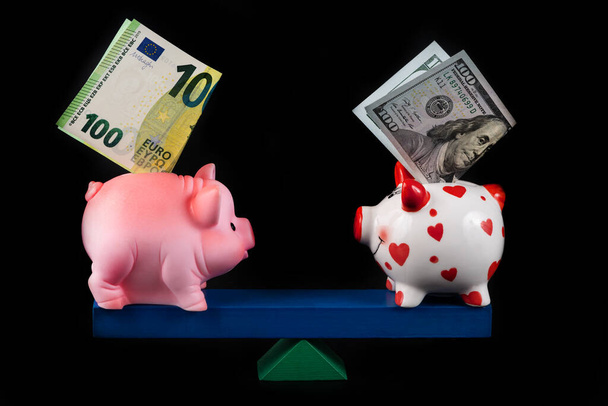 Varkensbanken met bankbiljetten van 100 euro en 100 Amerikaanse dollars in evenwicht op een balansbord op een zwarte achtergrond - Foto, afbeelding