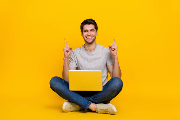 Full length photo of funny brunet millennial guy sit with laptop index up wear t-shirt jeans shoes isolated on yellow background. - Foto, immagini