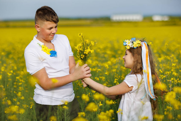Cheerful joyful carefree children: brother and sister in floral Ukrainian wreath with multi-colored ribbons sniff wild rapeseed flowers in bunches against backdrop of blooming yellow fields and sky - Фото, изображение
