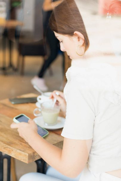 Kahve içen bir kız, kahve dükkanının arkasındaki cep telefonundan e-postaları kontrol eden genç bir kadın. - Fotoğraf, Görsel