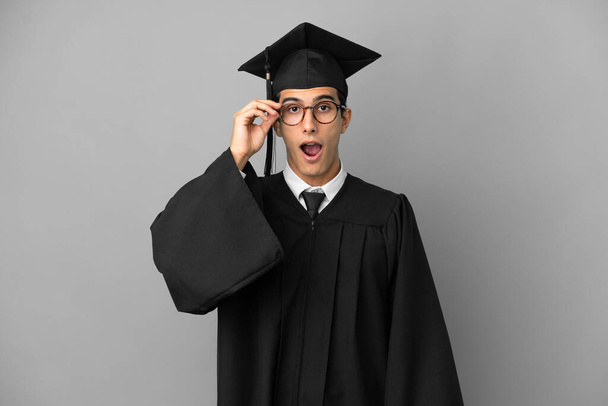 Young Argentinian university graduate isolated on grey background with glasses and surprised - Foto, afbeelding