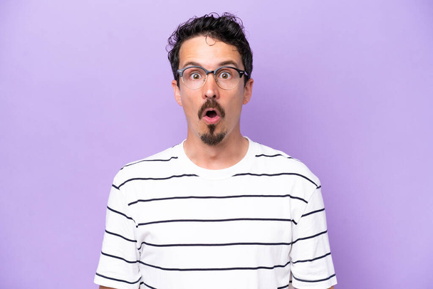 Joven hombre caucásico aislado sobre fondo púrpura con gafas y expresión sorprendida - Foto, imagen
