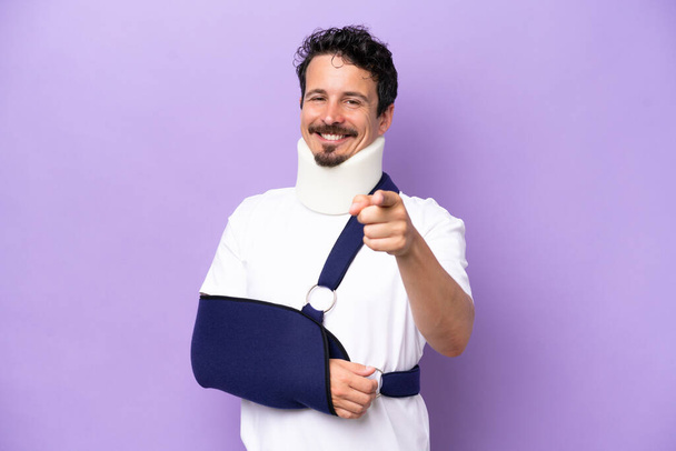 Young caucasian man wearing a sling and neck brace isolated on purple background pointing front with happy expression - 写真・画像