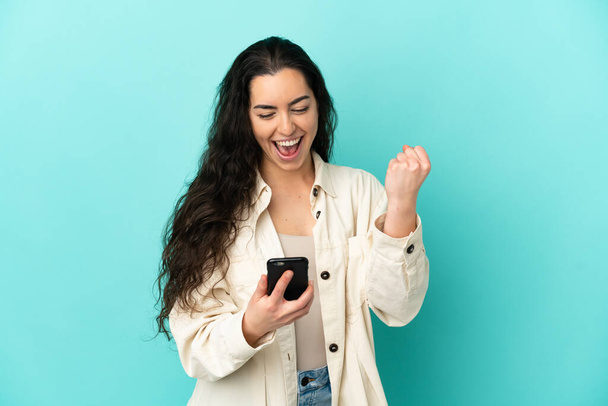 Young caucasian woman isolated on blue background with phone in victory position - Photo, Image