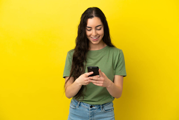 Young caucasian woman isolated on yellow background sending a message with the mobile - Zdjęcie, obraz