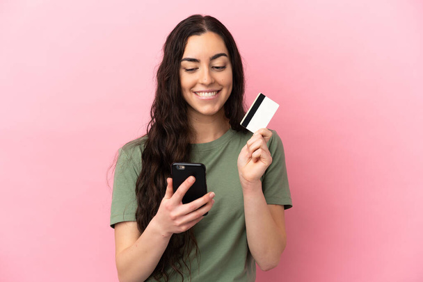 ピンクの背景に孤立した若い白人女性クレジットカードで携帯電話を購入しながら考える - 写真・画像