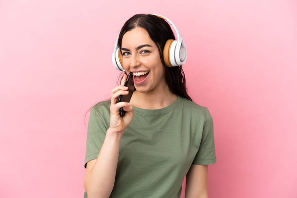 Junge kaukasische Frau isoliert auf rosa Hintergrund Musik hören mit einem Handy und singen - Foto, Bild