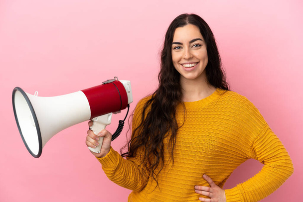 Young caucasian woman isolated on pink background holding a megaphone and smiling a lot - Fotografie, Obrázek