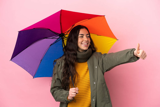 Elinde pembe arka planda izole edilmiş şemsiyesi olan genç bir kadın baş parmak hareketi yapıyor. - Fotoğraf, Görsel