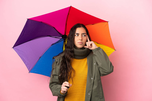 Молода жінка тримає парасольку ізольовано на рожевому фоні думаючи про ідею
 - Фото, зображення