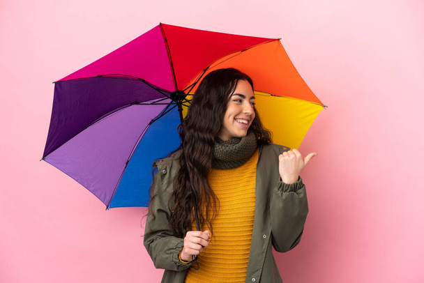 Молода жінка тримає парасольку ізольовано на рожевому тлі, вказуючи на бік, щоб представити продукт
 - Фото, зображення